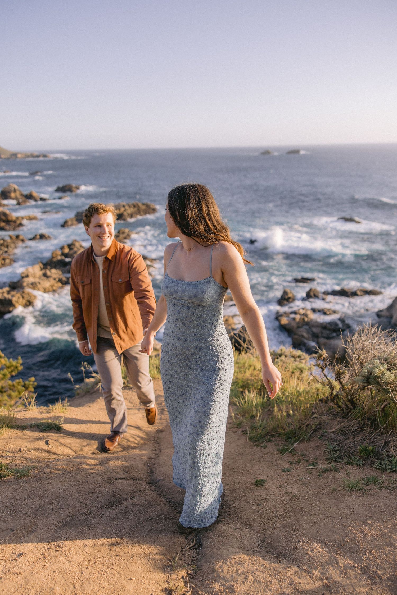 Couple walking along scenic coastal trail with crashing waves and rocky shoreline during golden hour at sunset.