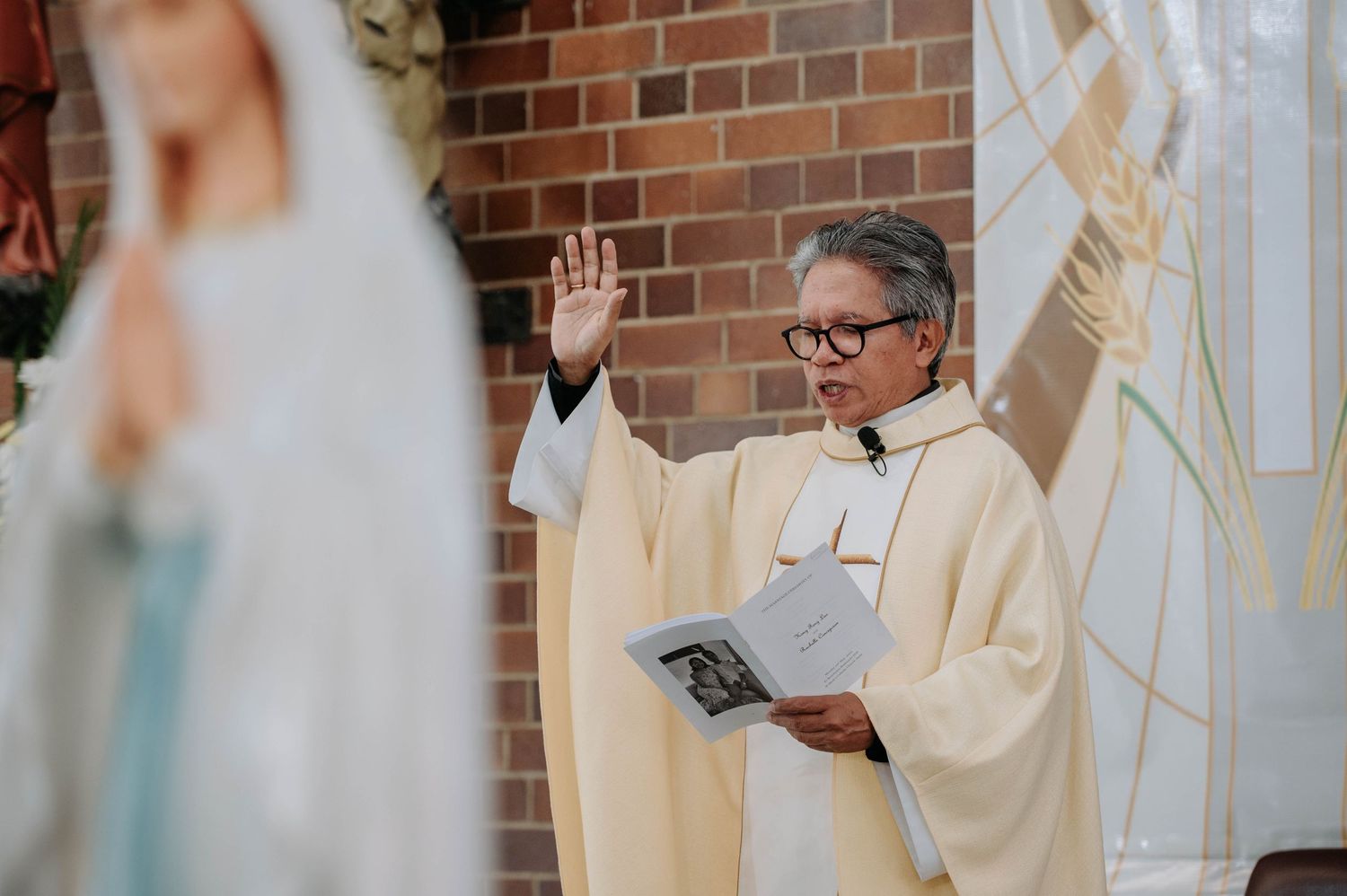 Faith leader in cream-colored robes raises hands while holding a book during religious ceremony at brick church.