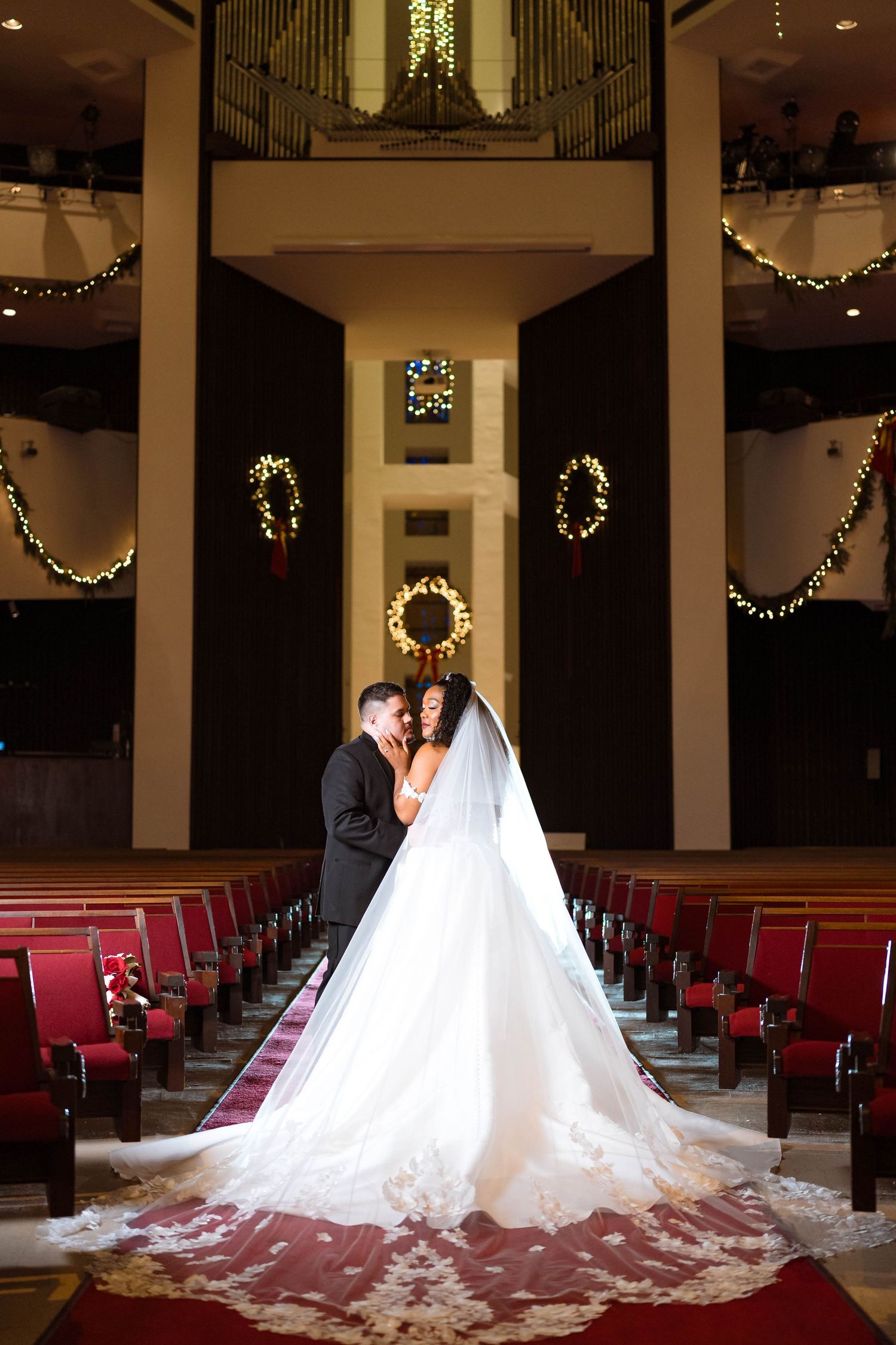 A wedding couple stands in a grand church aisle with ornate organ pipes and holiday decorations along the balconies.