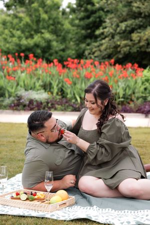 Couple enjoys romantic picnic on blanket with charcuterie board and wine in garden with red tulips blooming.