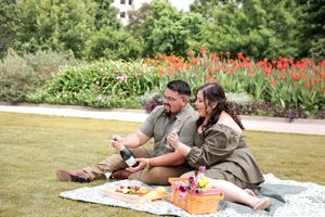 A couple enjoys a romantic summer picnic on a blanket in a garden with red flowers blooming in the background.