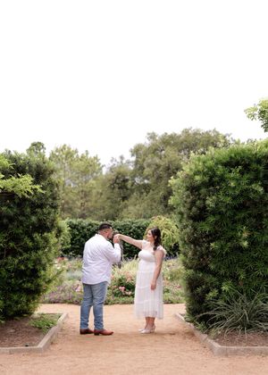 Two people dancing together on a garden path surrounded by lush green hedges and trees on a sunny day.