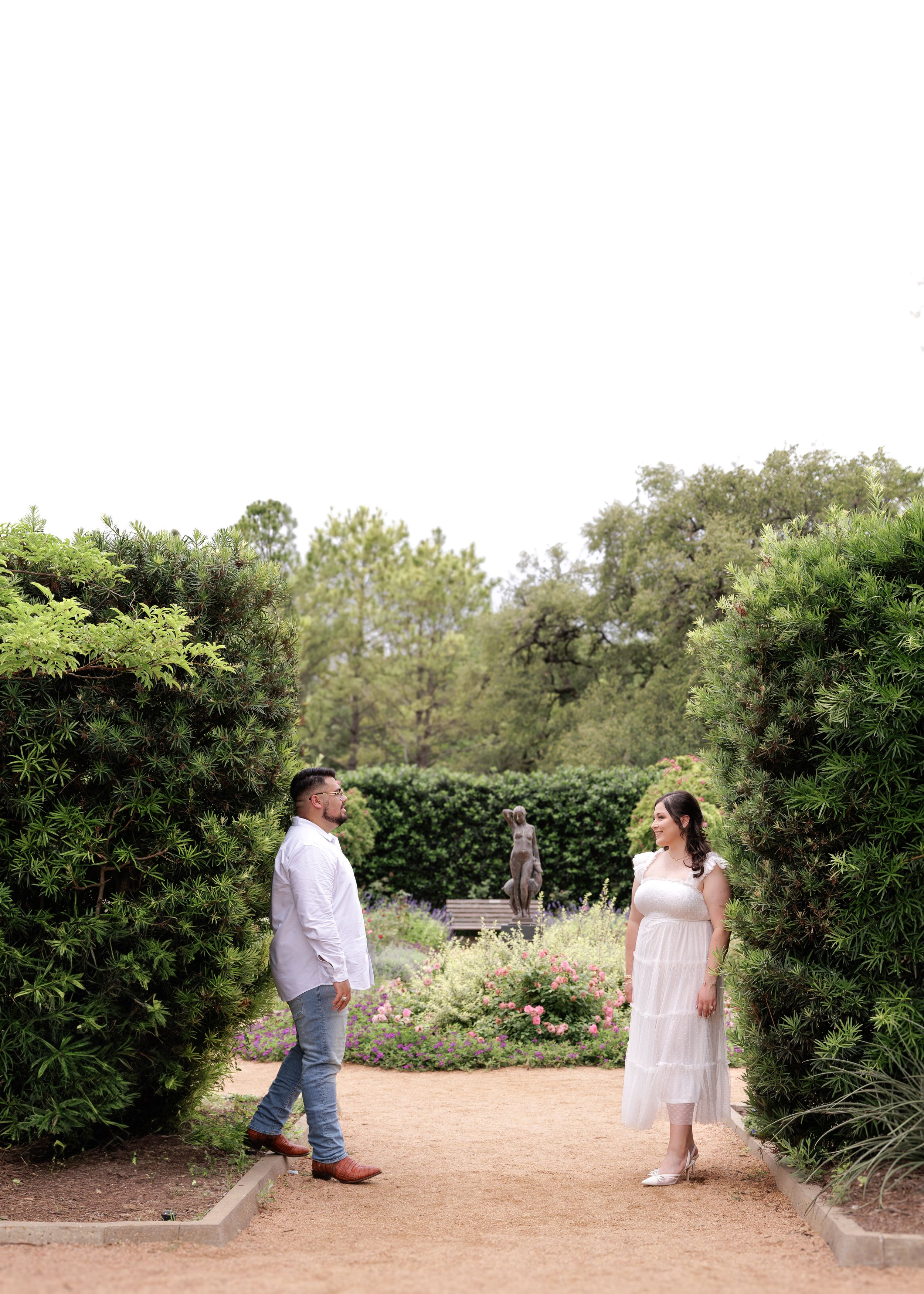 A couple stands facing each other on a dirt path in a garden with flowering shrubs and hedges on a bright summer day.
