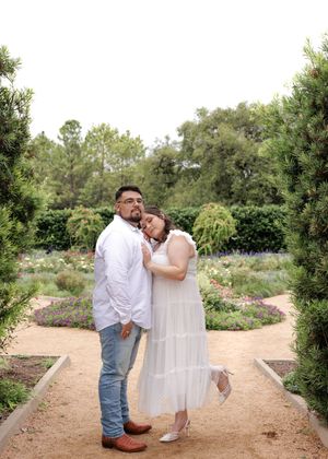 A couple embraces on a garden path surrounded by lush greenery and flowering plants on a sunny day.