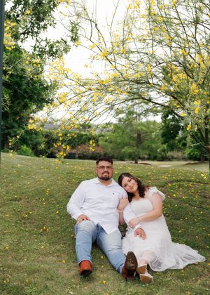 A couple sits together on grassy lawn beneath blooming yellow spring trees in an outdoor garden setting.