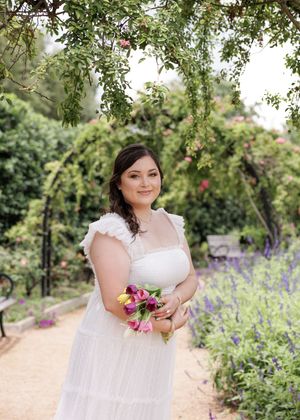 Person in white lace wedding dress holding pink flowers stands on garden path beneath overhanging tree branches.