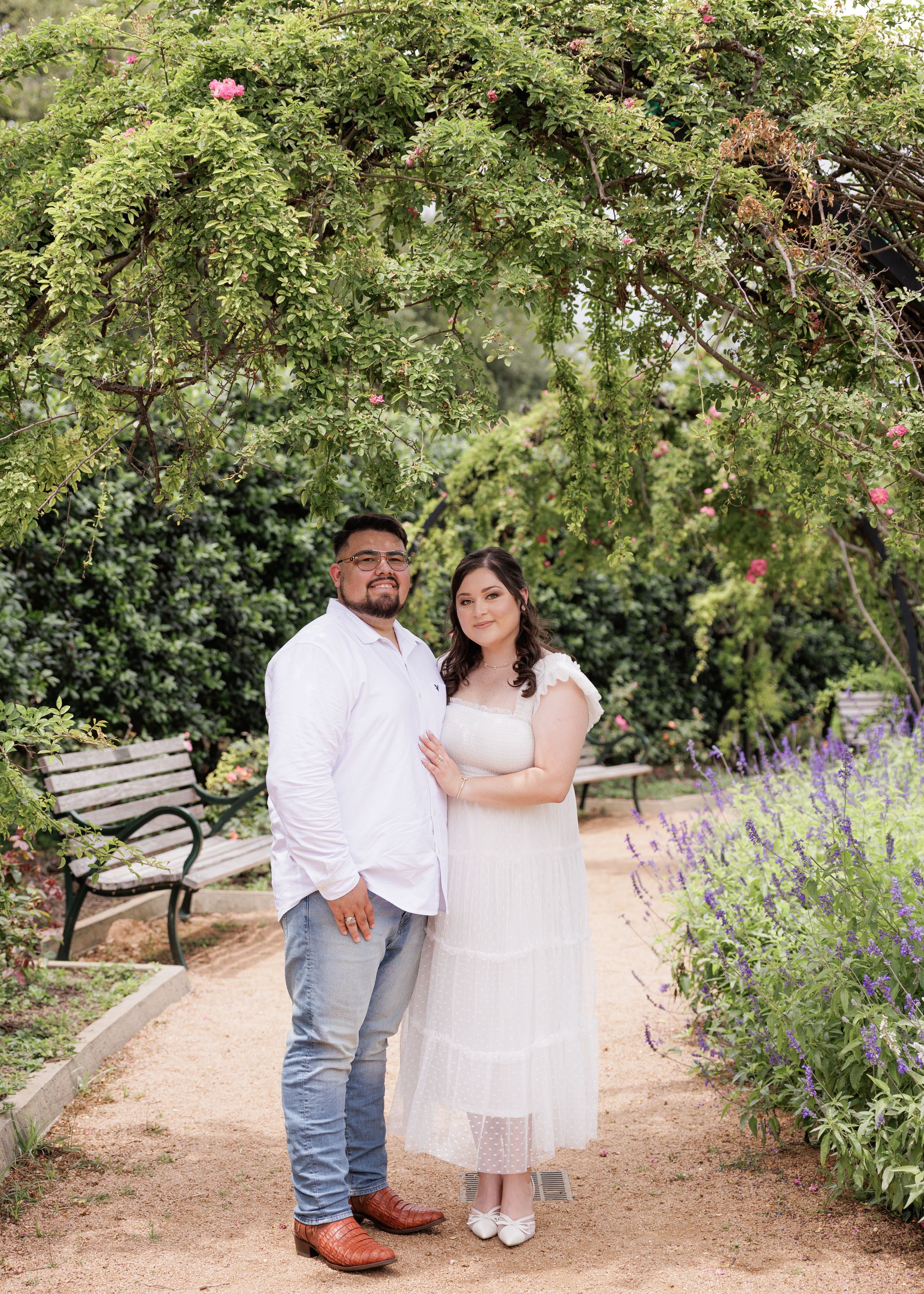 Couple poses together under a lush garden archway with blooming flowers and greenery along a gravel path.