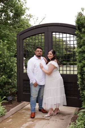 A couple poses together in front of a black arched garden gate surrounded by greenery.