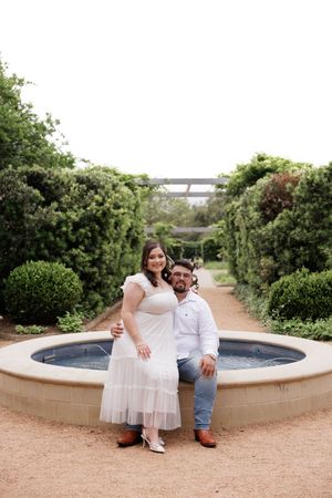 A couple poses together on a garden path next to an elegant stone fountain surrounded by lush greenery and hedges.