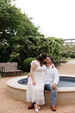 A couple in white dress and casual attire share an intimate moment while sitting by a circular fountain in a garden setting.