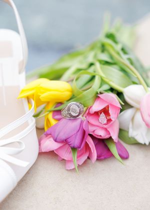 A diamond engagement ring rests elegantly on colorful spring tulips with pink, yellow and white petals beside white fabric.