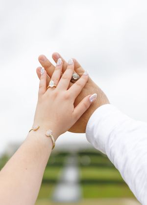 Close-up of hands with wedding rings and manicured nails touching against blurred outdoor background.