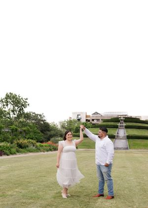 A couple in wedding attire dance together on a grassy lawn with a lighthouse and landscaped garden in the background.