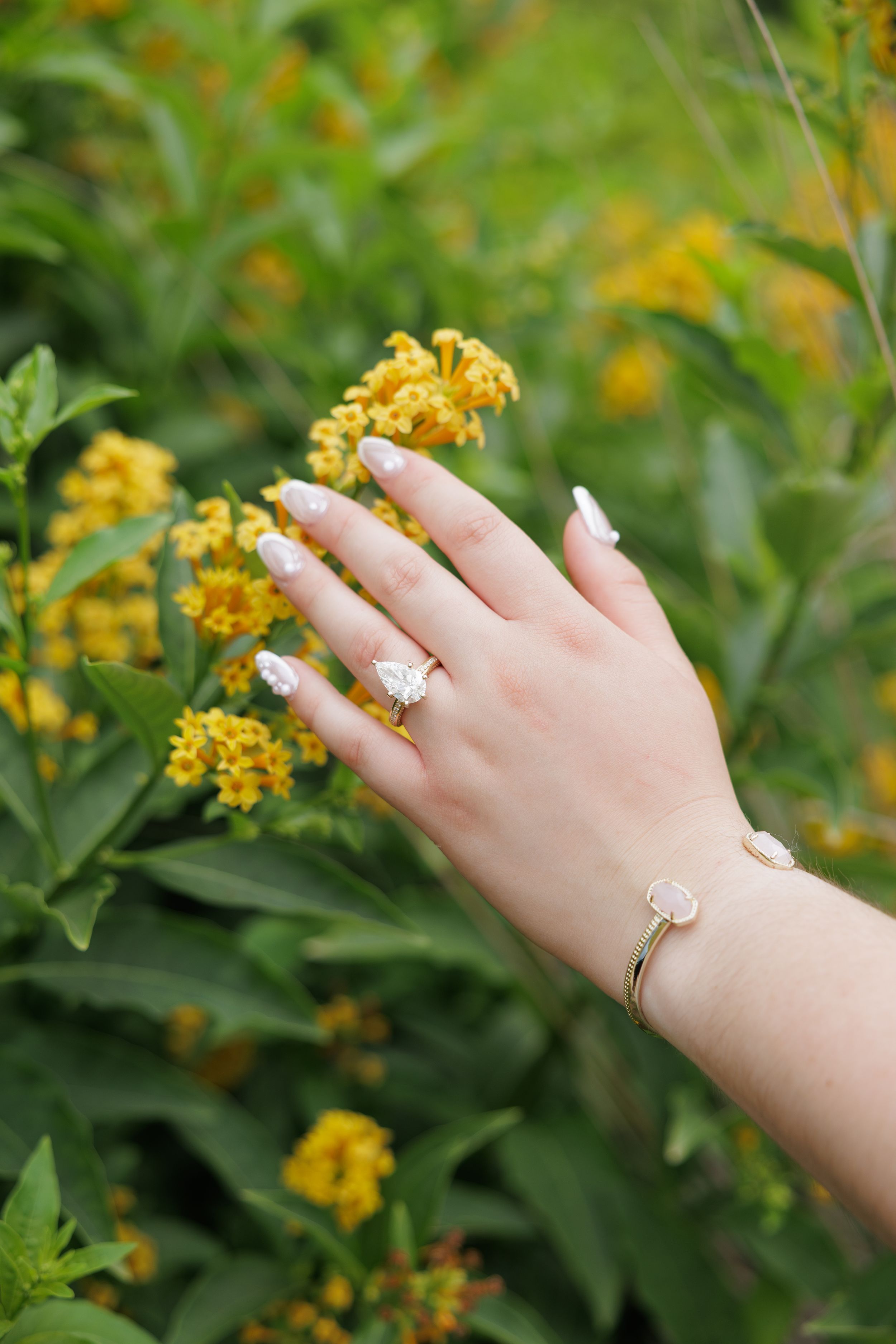 A delicate hand wearing a gold ring gently touches yellow flowers against a green garden background.