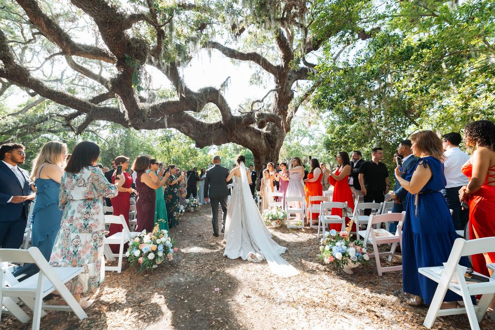 An outdoor wedding ceremony takes place under a large oak tree with guests seated on white chairs in a garden setting.