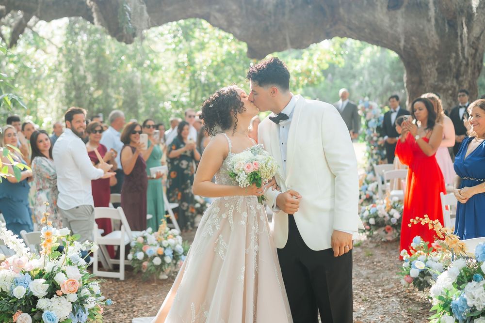 Couple shares romantic moment at outdoor wedding ceremony under oak trees with guests and floral arrangements in background.