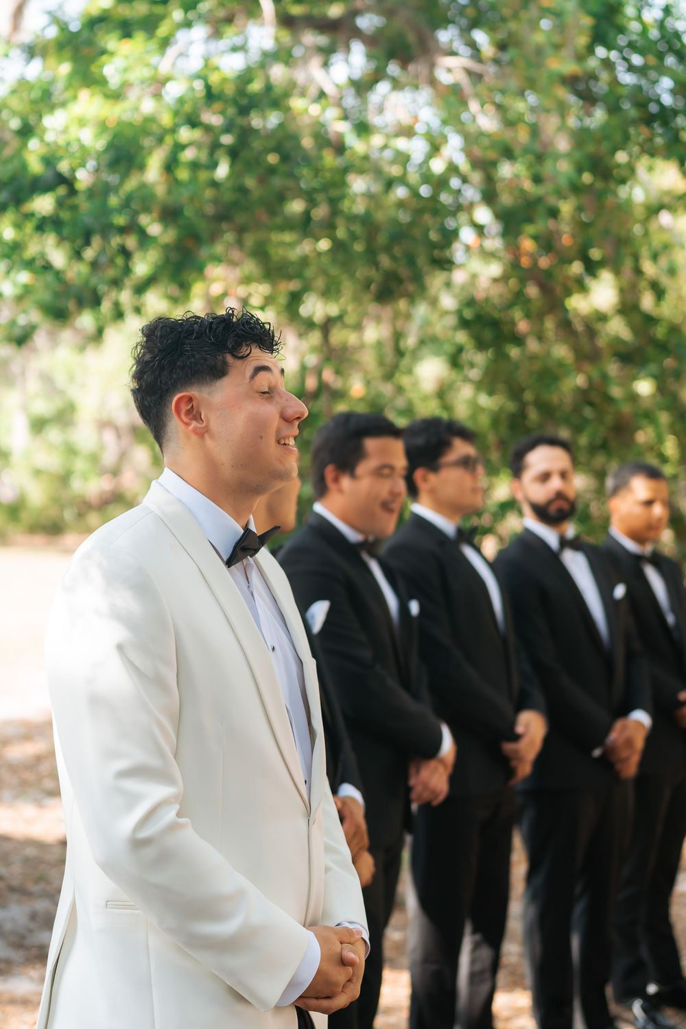 Groomsmen lineup at outdoor wedding with groom in white tuxedo jacket and others in classic black suits.
