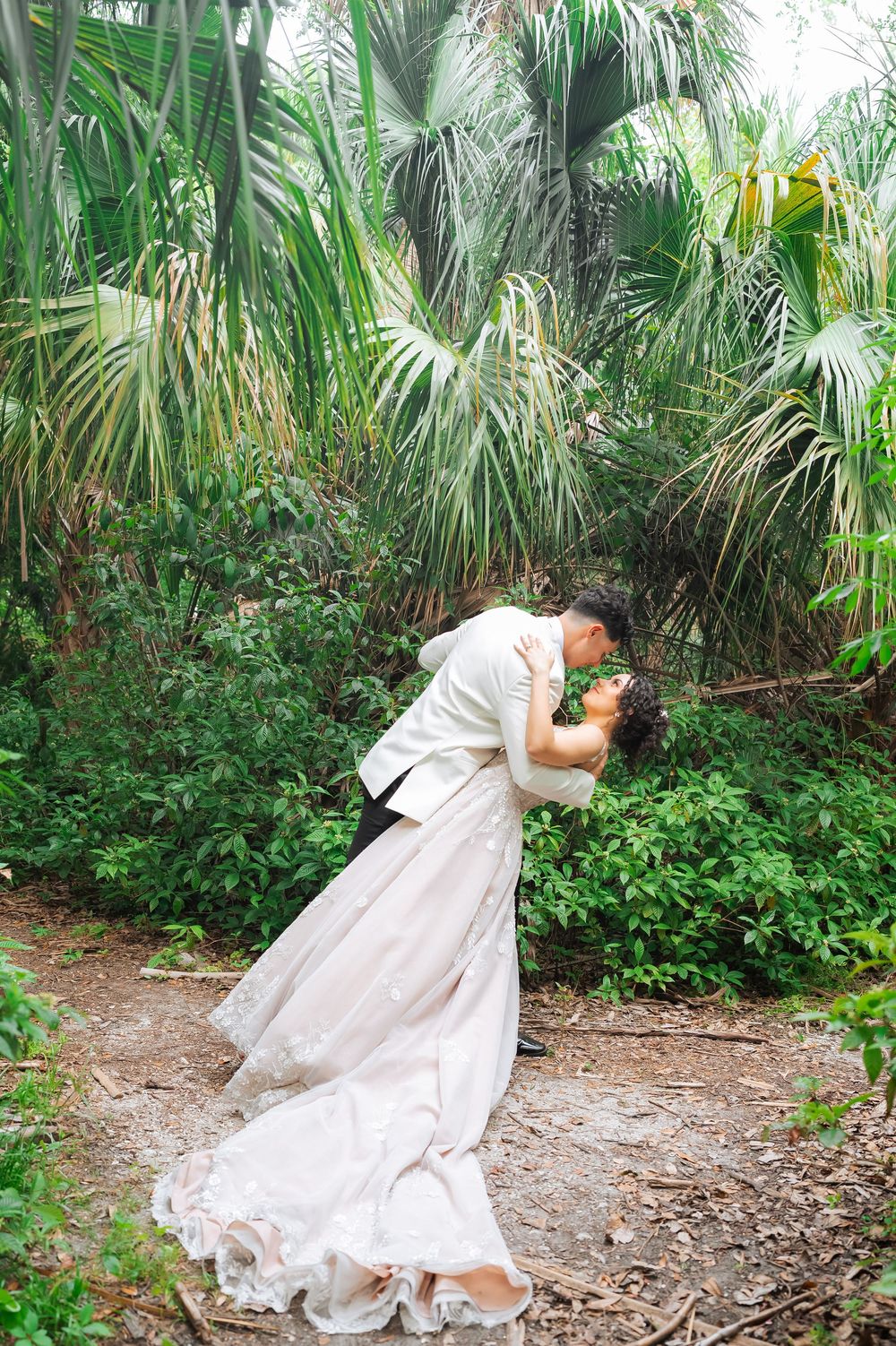 A romantic couple in formal attire share a romantic moment amongst tropical palm trees and lush greenery.