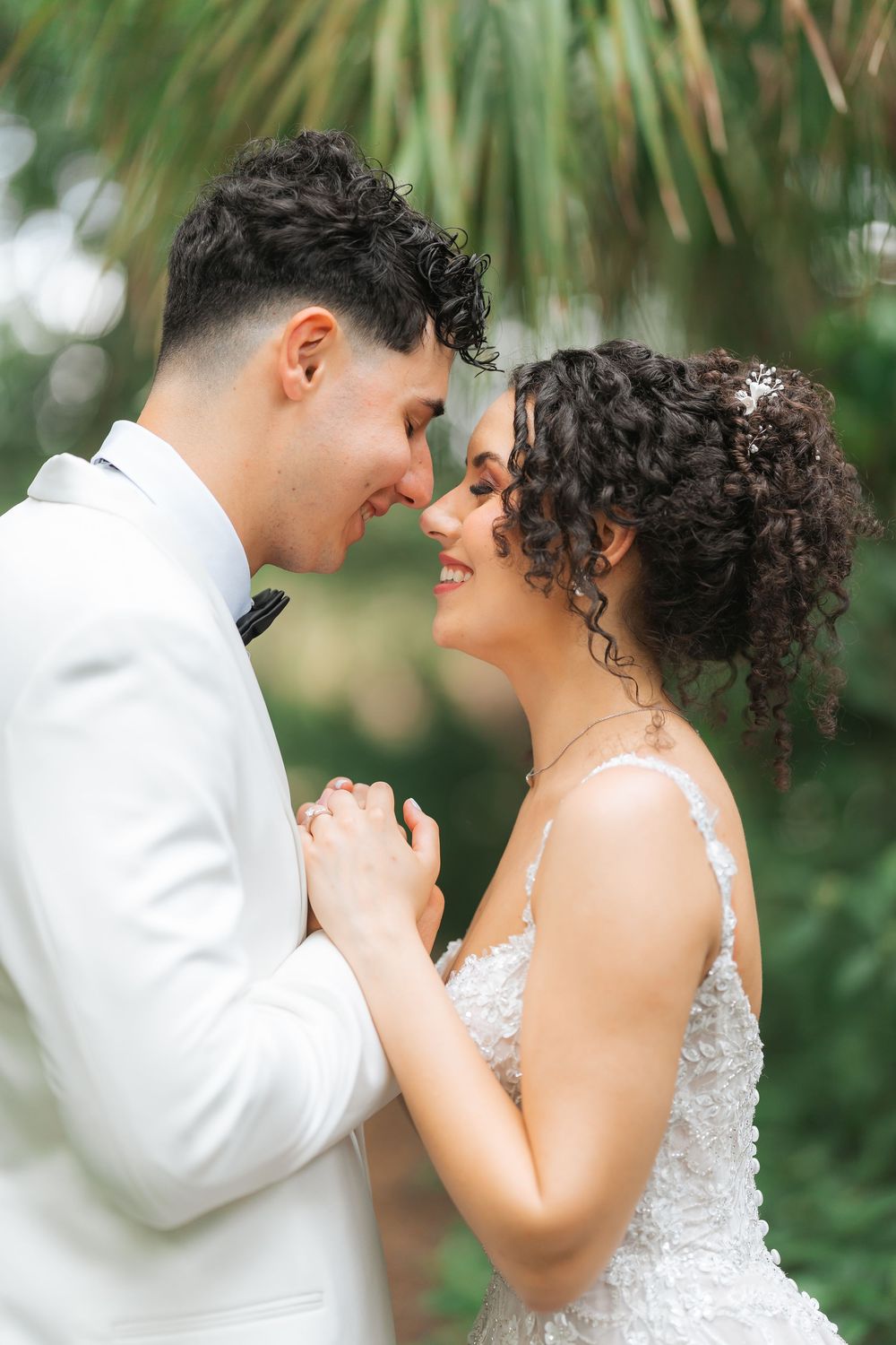A romantic wedding couple holds hands and leans in close together against a backdrop of tropical palm fronds.