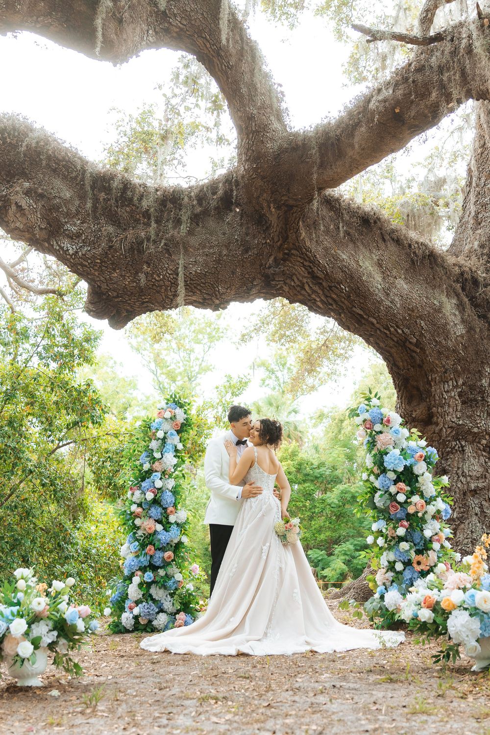 A romantic wedding scene under a majestic oak tree with blue and white floral arrangements framing a couple embracing.