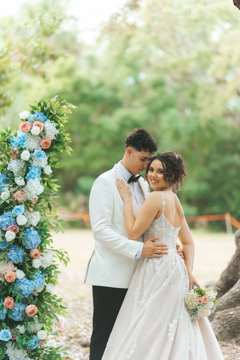 A romantic wedding couple embraces by a floral arch with blue and peach blooms in an outdoor garden setting.