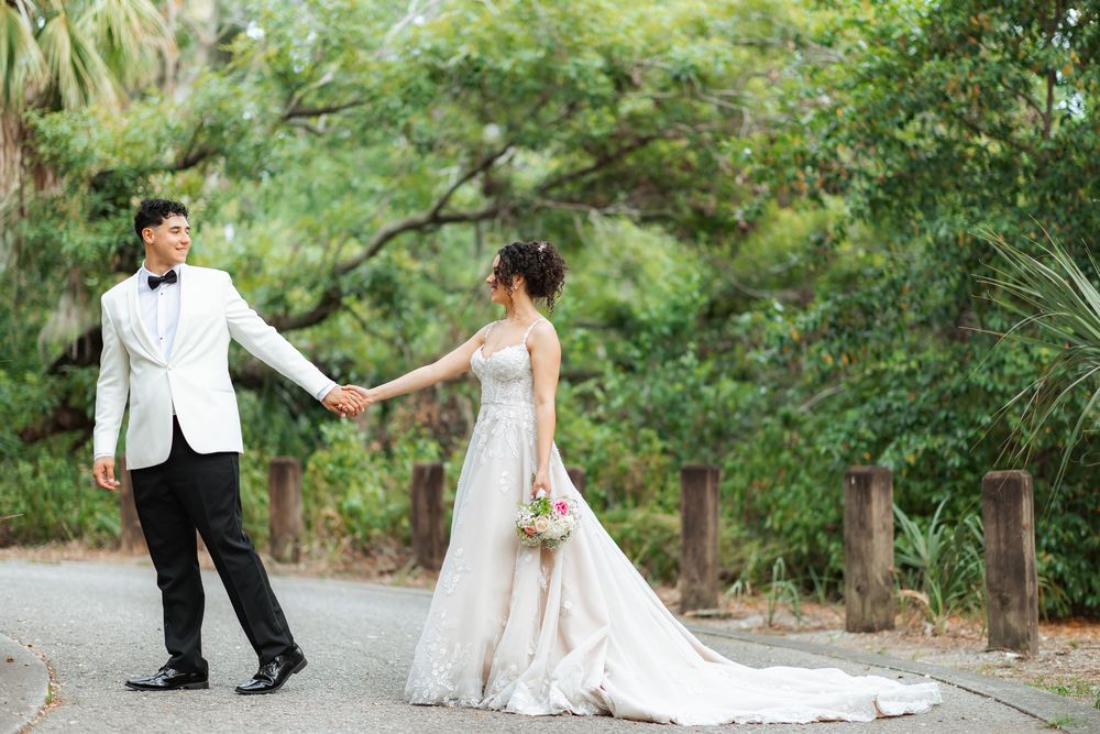 A couple in wedding attire hold hands while walking on a garden path surrounded by lush greenery.