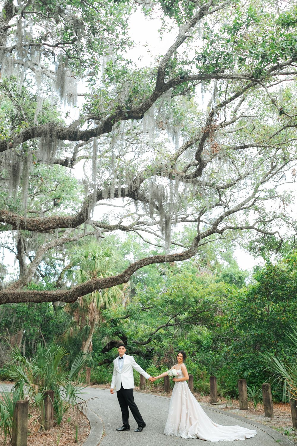 Beautiful wedding couple dances on pathway under majestic Spanish moss covered oak trees in a romantic garden setting.