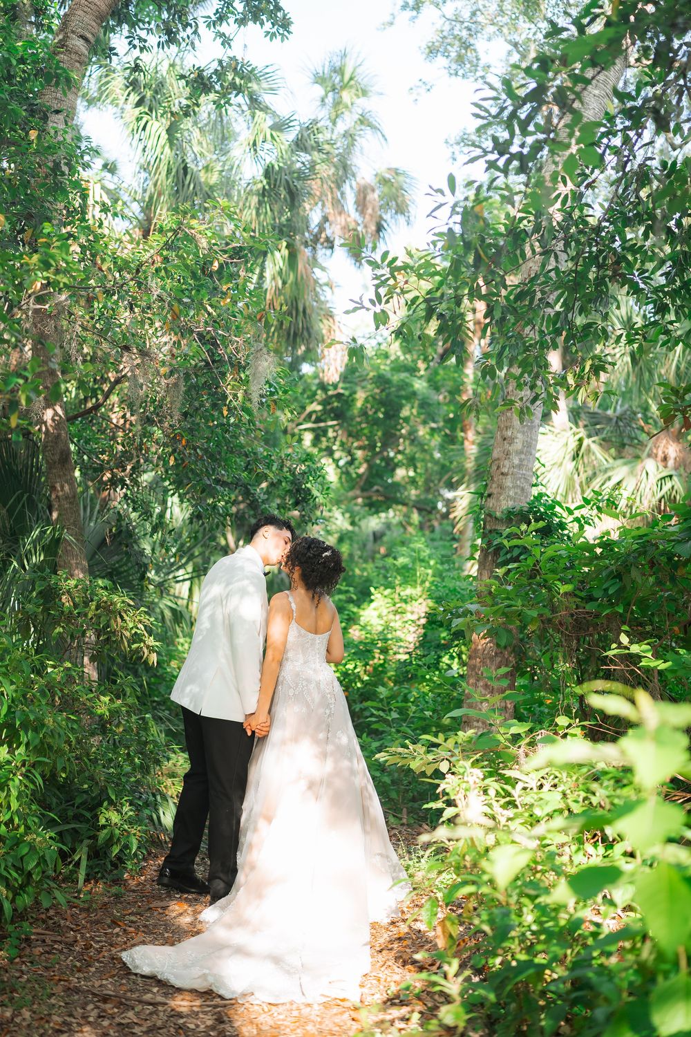 A couple shares an intimate moment on a garden path surrounded by lush tropical foliage and palm trees.