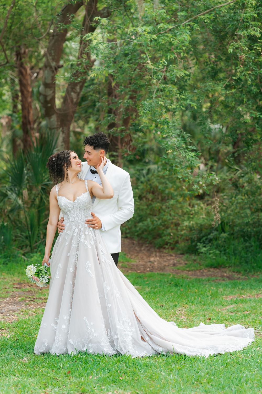 A romantic wedding couple poses together in a lush garden setting, with the bride wearing a flowing lace gown with long train.