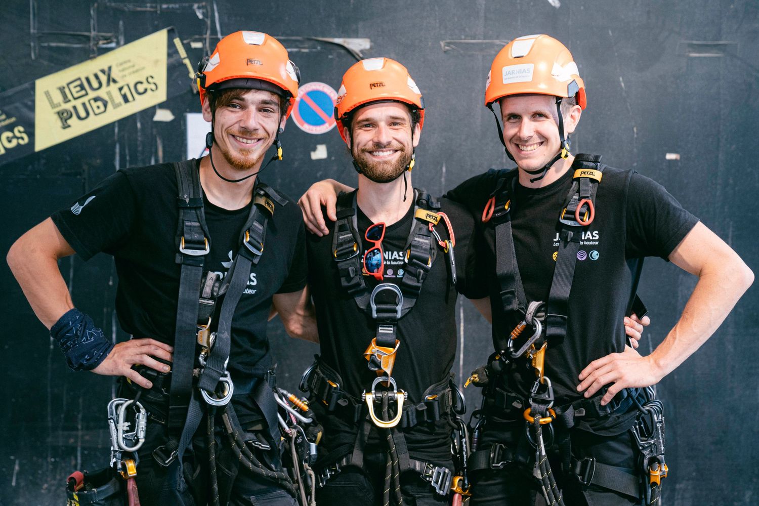 Three workers wearing orange hard hats and black safety harnesses smile while posing against dark wall.