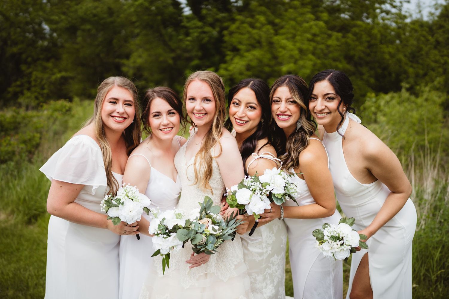 A joyful bridal party in white dresses poses together outdoors holding white and green floral bouquets.