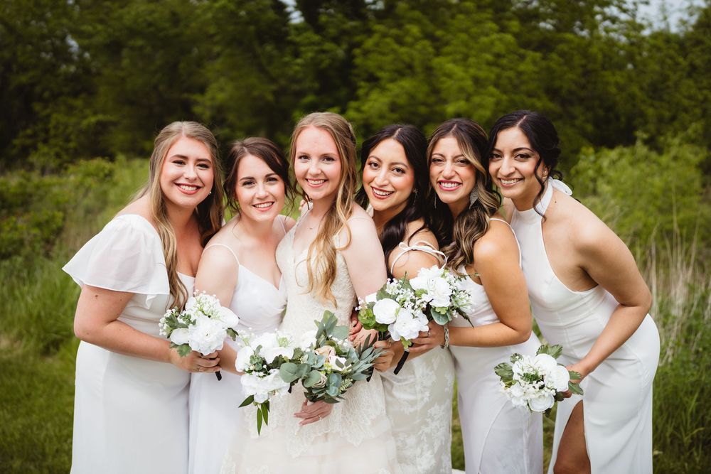 A joyful bridal party in white dresses poses together outdoors holding white and green floral bouquets.