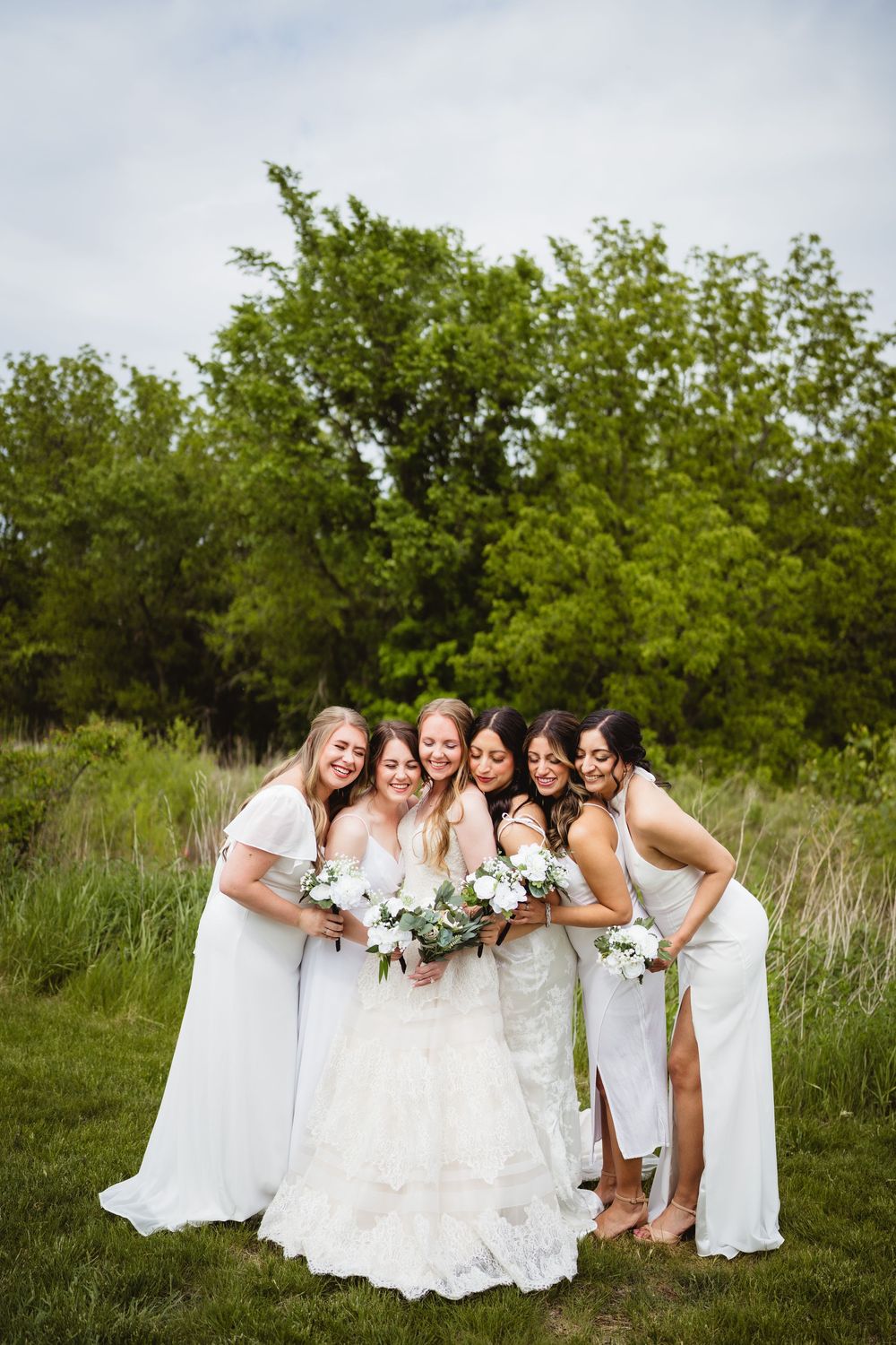 A bridal party in white dresses poses together on grass with trees in the background, holding white floral bouquets.