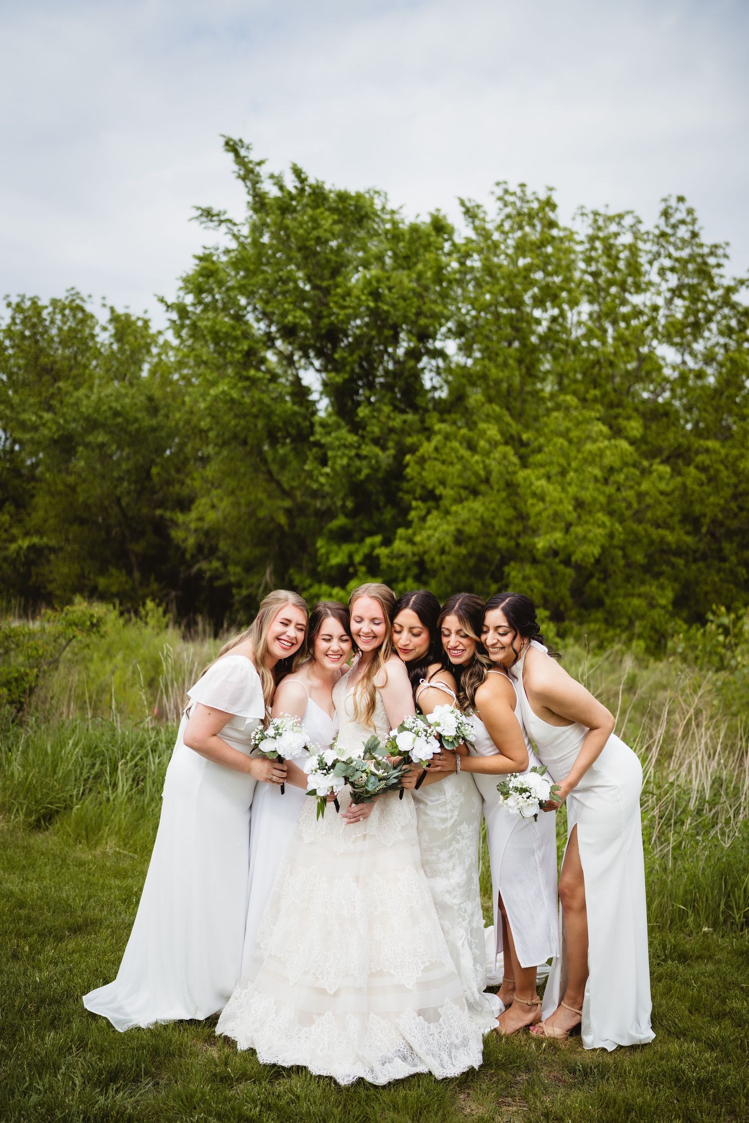 A bridal party in white dresses poses together on grass with trees in the background, holding white floral bouquets.