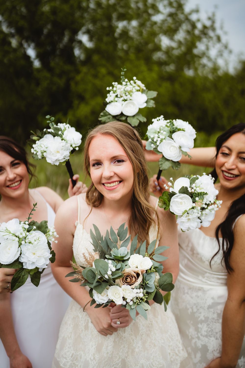 A close up of a bridal party sharing laughter while holding white flower bouquets outdoors.