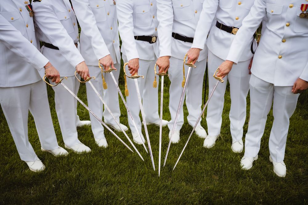 Row of people wearing coordinated white uniforms and holding sabers standing on grass in military formation.