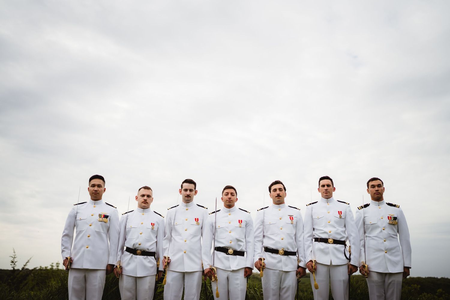 A row of military cadets in white uniforms and belts stand together outdoors against a cloudy sky.