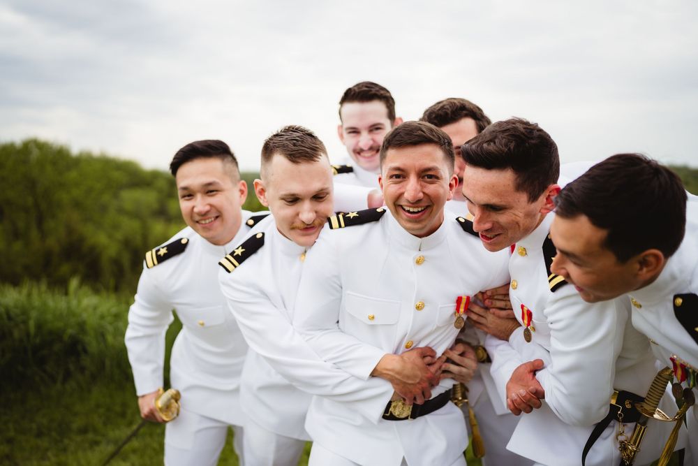 A group of military officers in white uniforms laugh together while standing in a grassy outdoor setting.