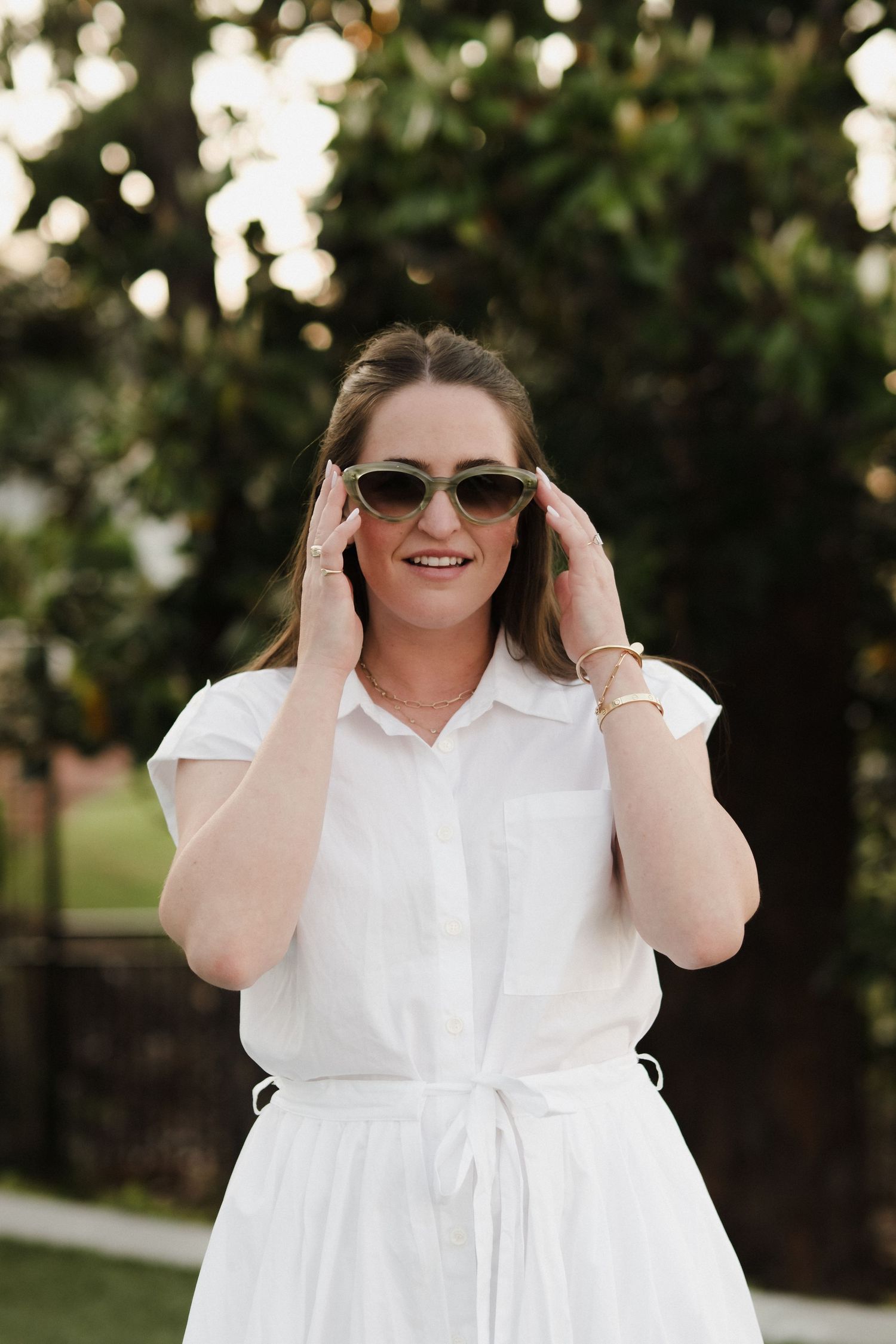 Woman in white dress adjusting sunglasses outdoors in natural lighting.