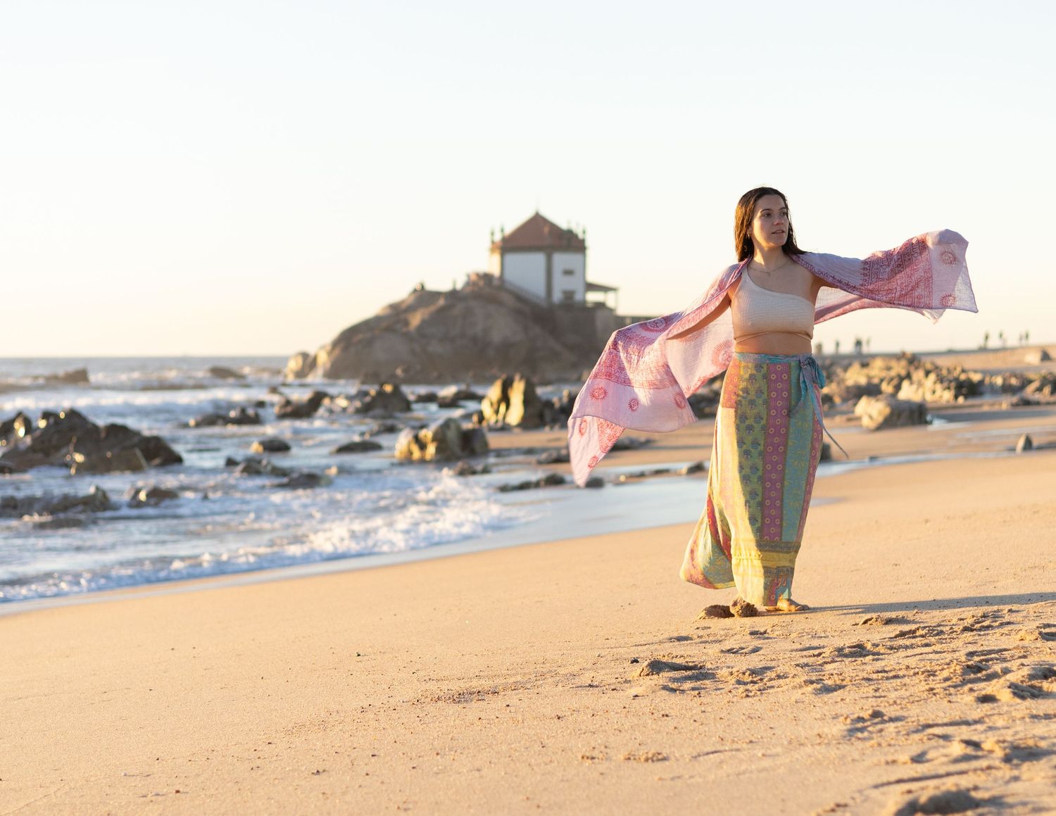 Woman in flowing clothes dancing on beach at sunset near lighthouse.