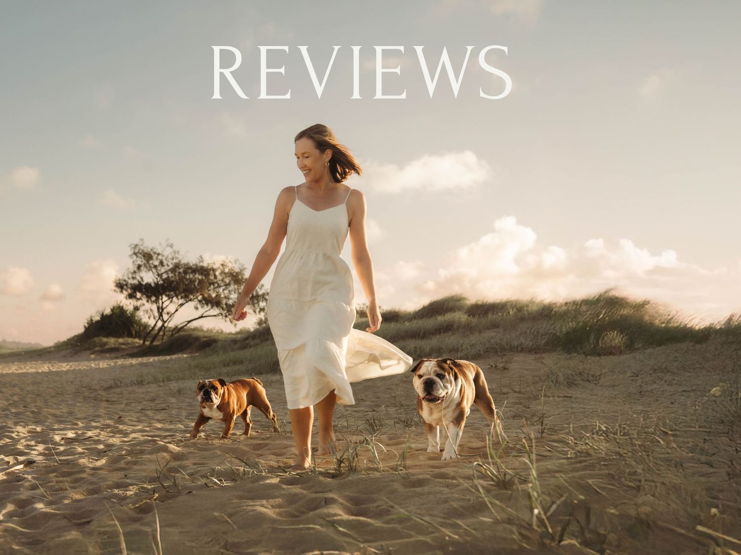 Two dogs walking with someone in a white dress on a sandy beach during sunset.