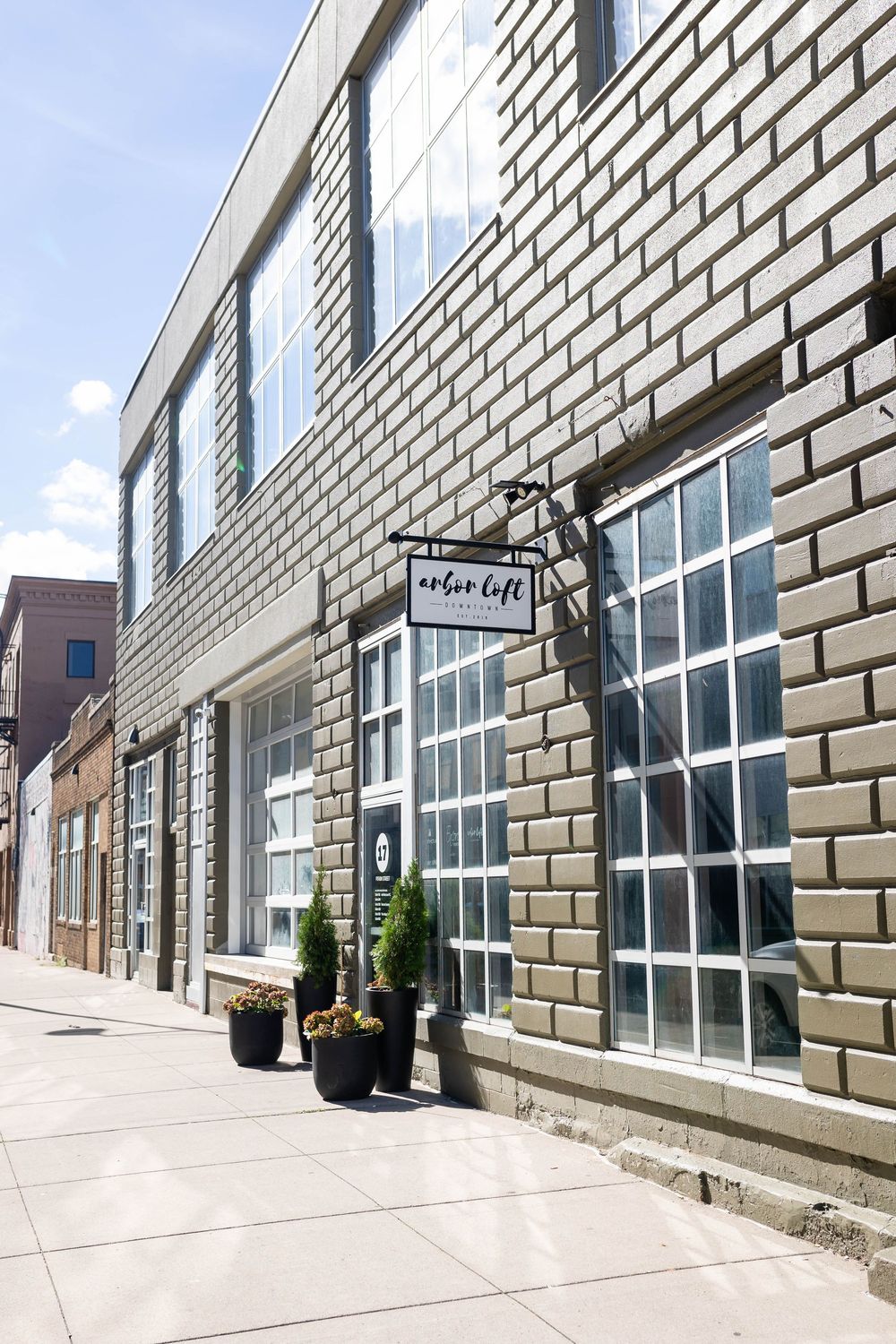 Modern brick building facade with large windows, black signage, and decorative potted plants along a sunny sidewalk.
