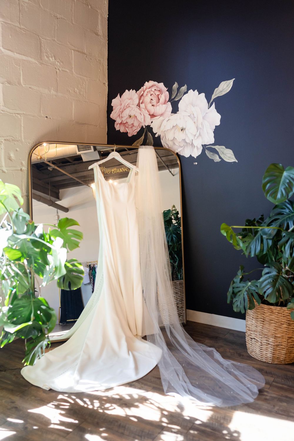 Elegant wedding dress hanging on gold-framed mirror with pink and white floral arrangement and potted plants in bridal suite.