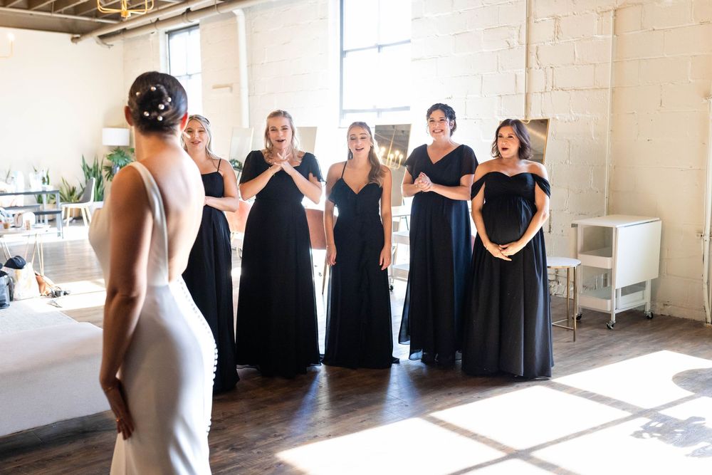 A person in a white dress shows their outfit to a group in matching navy gowns in a sunlit room.