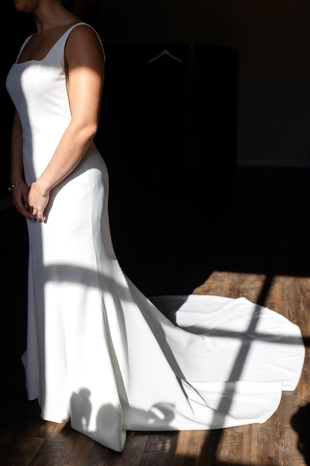 Elegant white wedding dress with flowing train casts dramatic shadows on wooden floor in dramatic lighting.