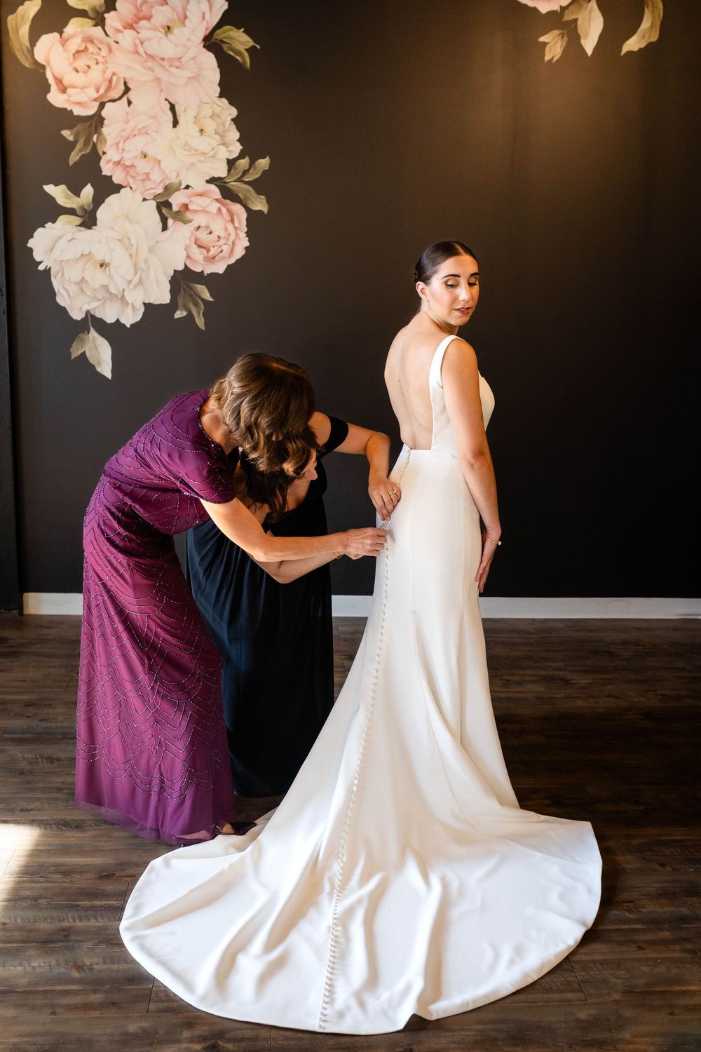A woman in a purple dress helps adjust an elegant white wedding gown with a long train against a dark floral wall.