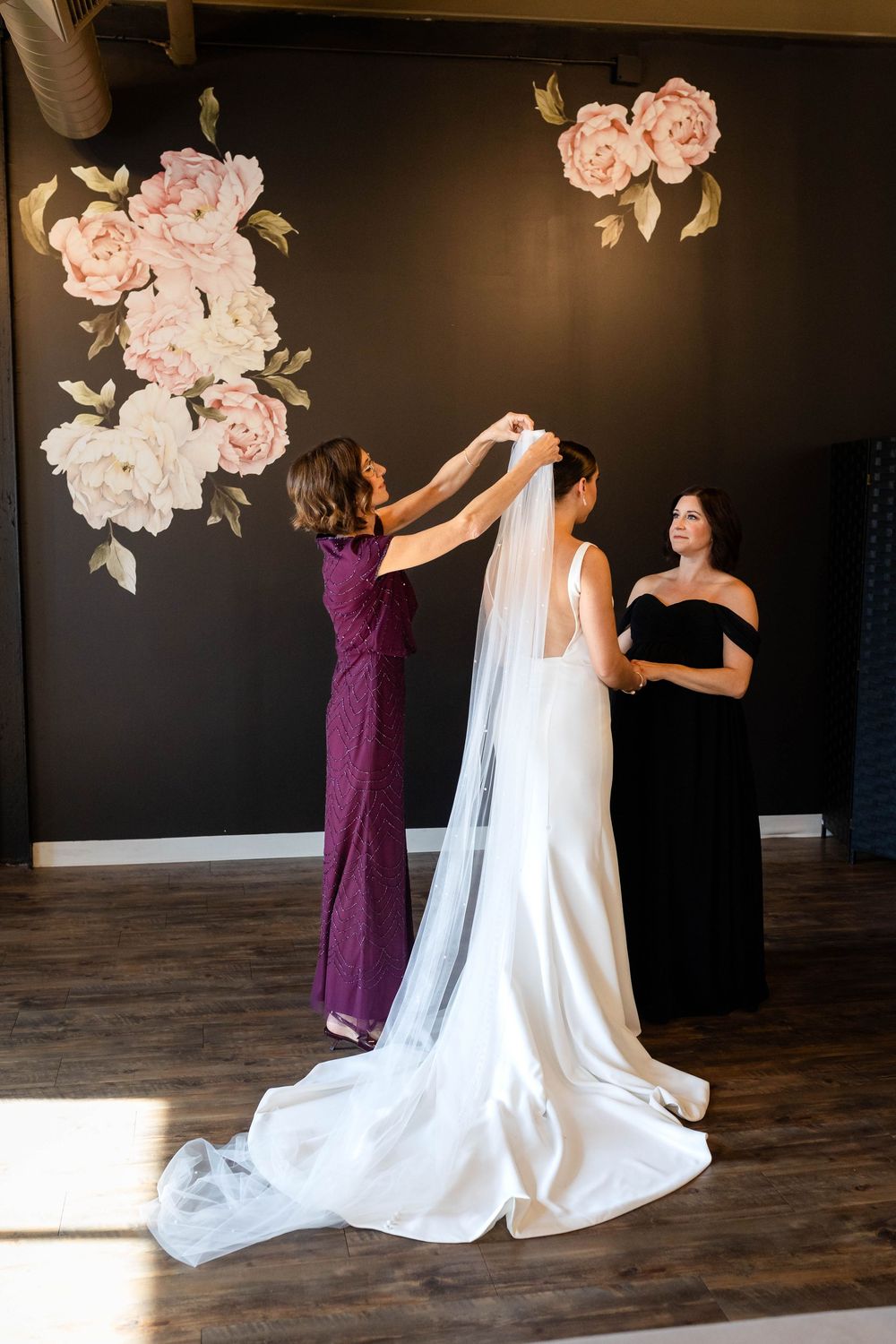 Wedding preparation scene with bridesmaids helping adjust a long white veil against a dark wall with pink floral accents.