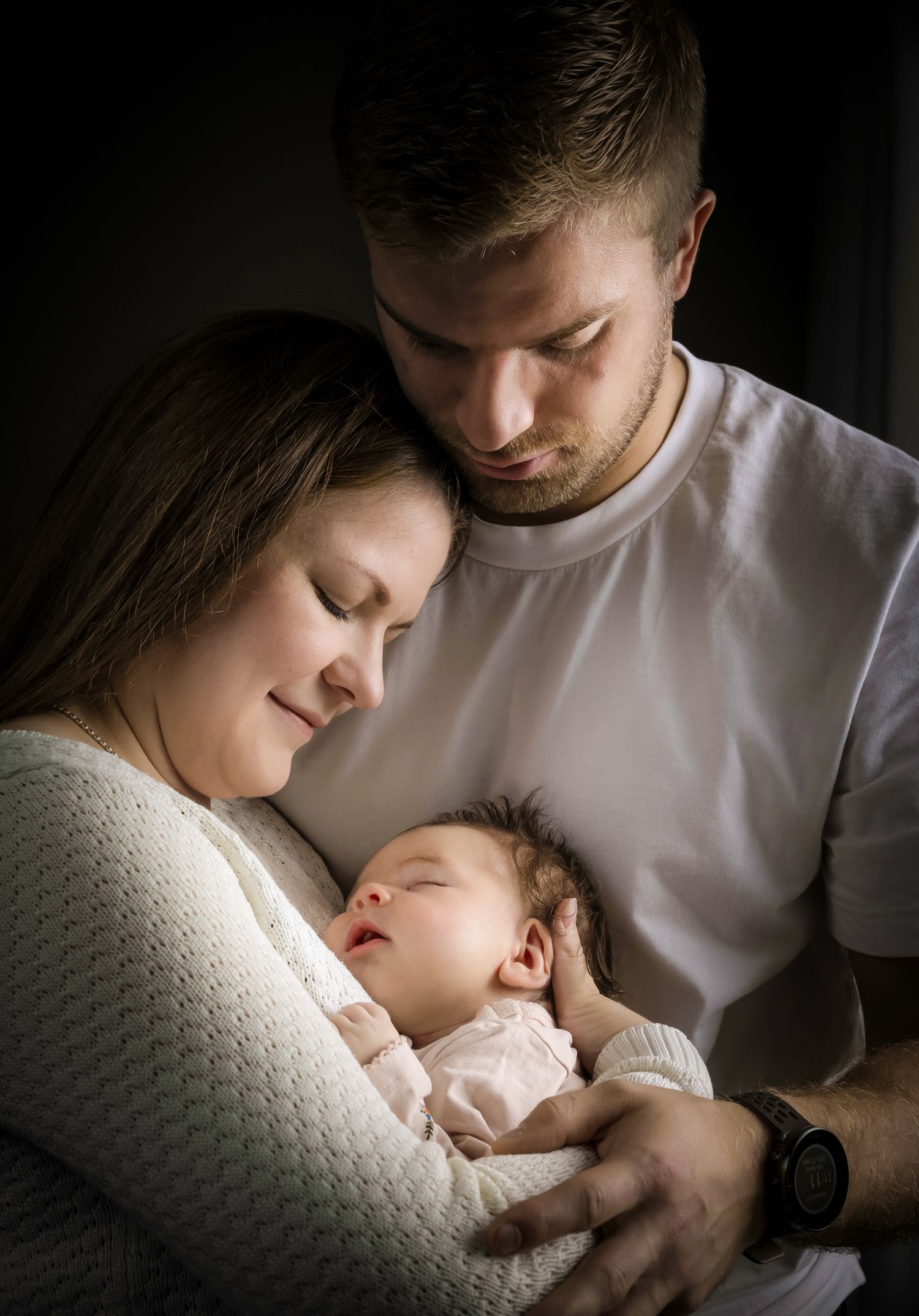 New parents tenderly cradling their sleeping newborn baby, sharing a quiet intimate family moment in soft natural light.