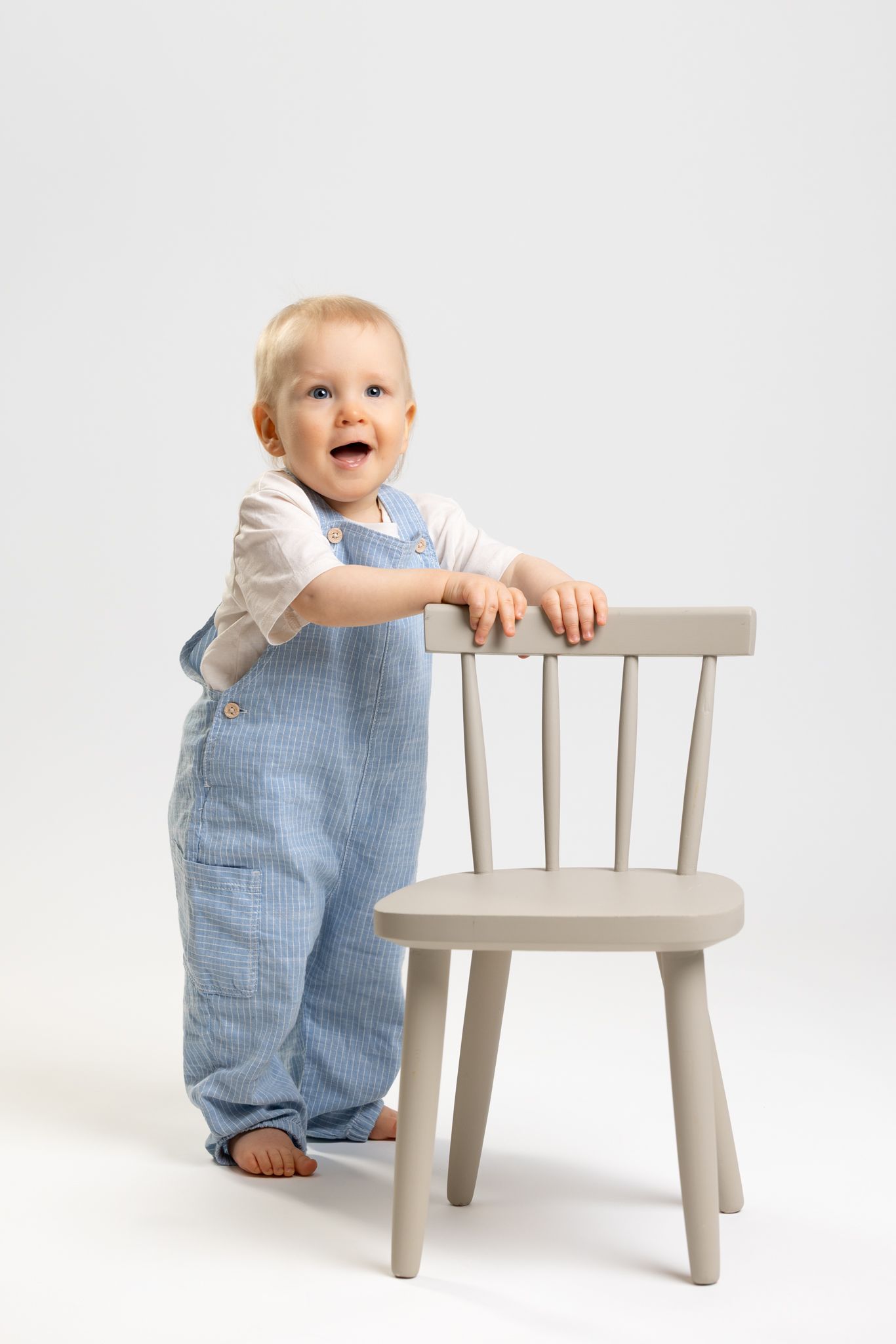 Happy toddler in blue overalls standing beside a wooden chair, smiling brightly against a white background.