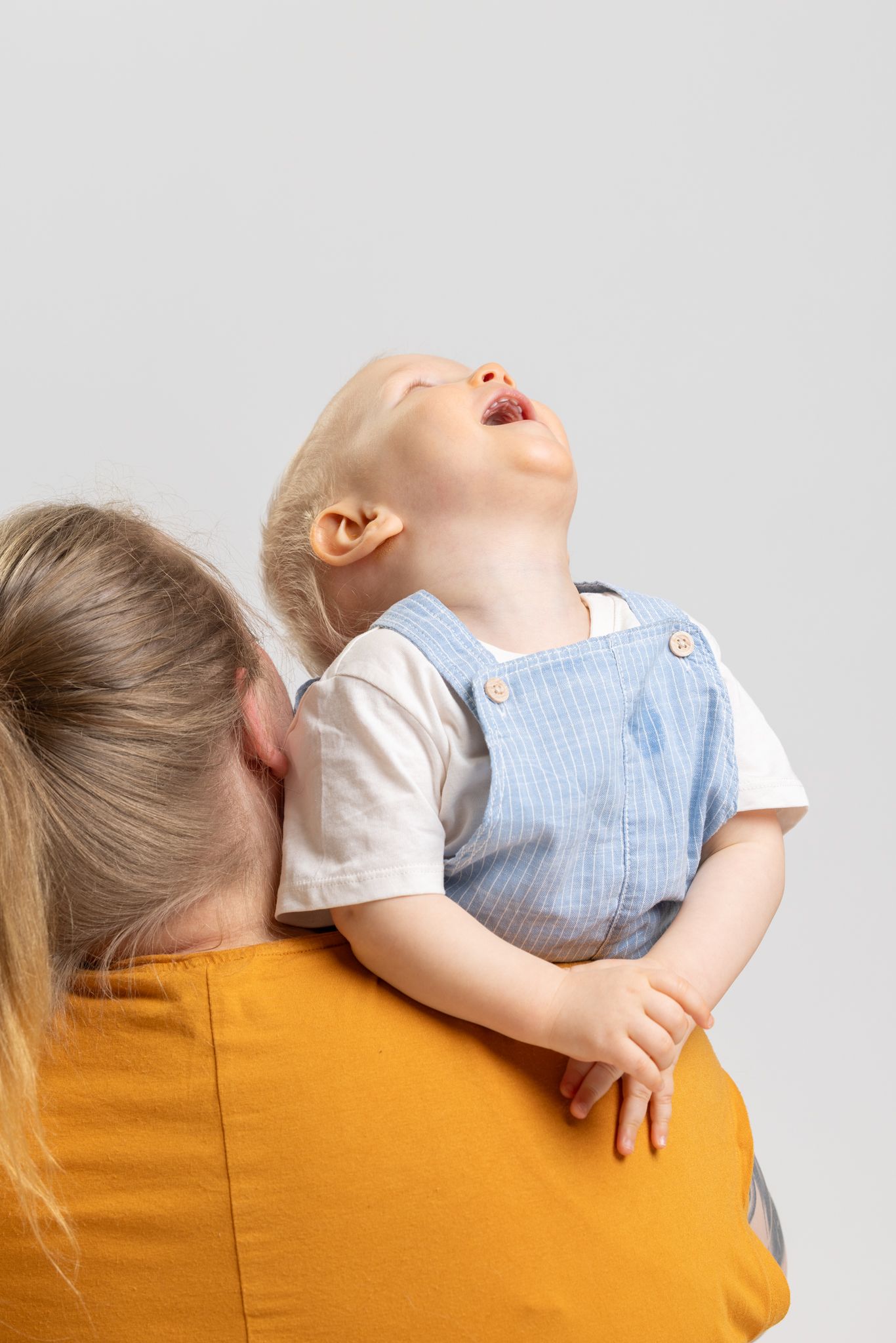 A mother in a yellow shirt holds a laughing toddler in blue overalls against a light gray background.